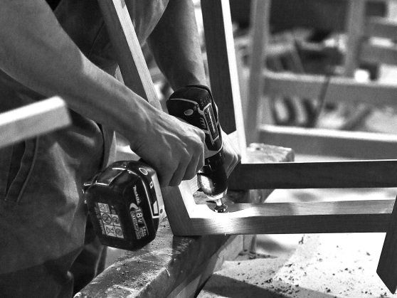 Ein Schwarzweißfoto einer Person, die mit einem Bohrer ein Stück Holz bearbeitet.