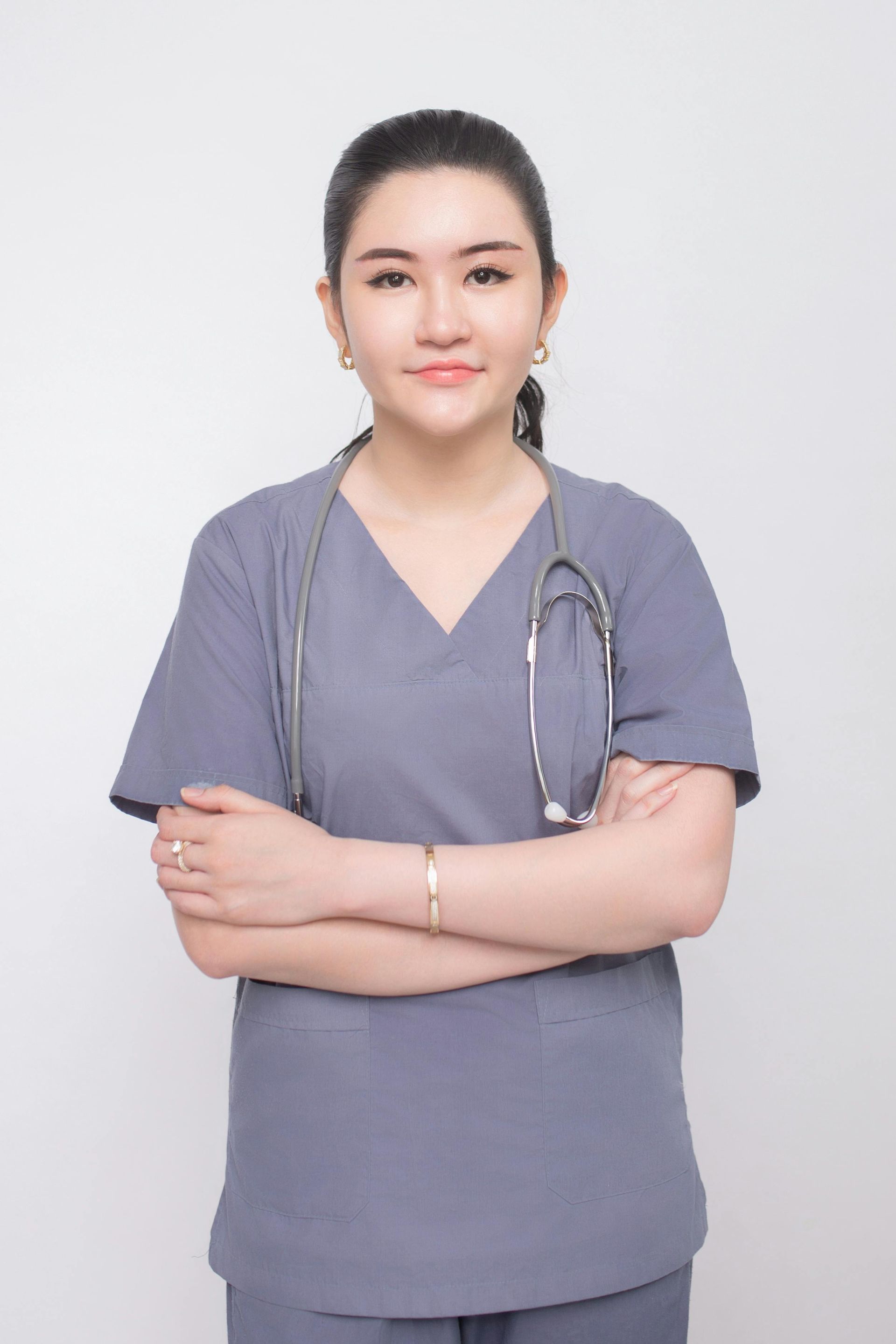 Woman in gray scrubs with a stethoscope, arms crossed, smiling, against a white background.