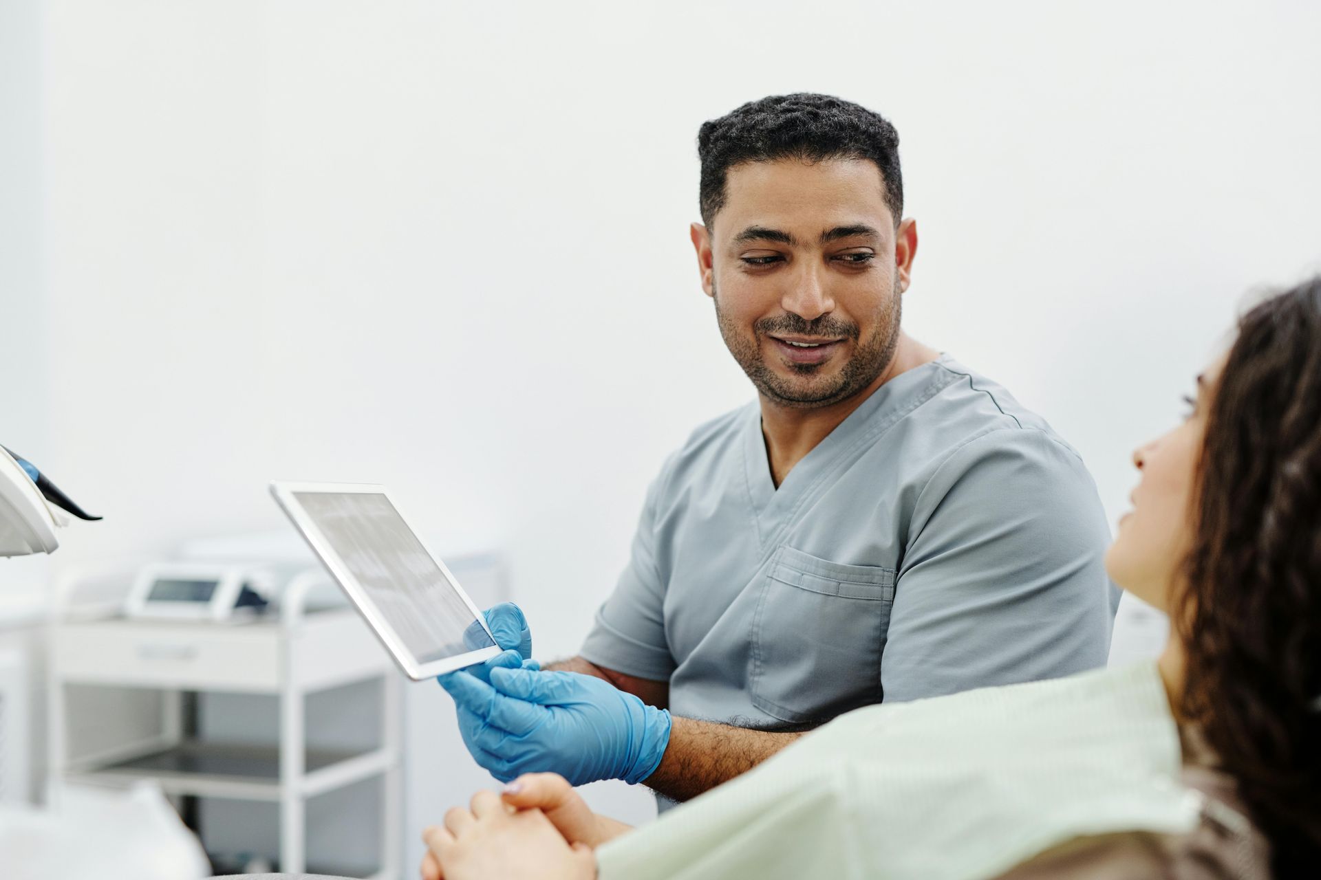 Dentist in scrubs and gloves showing a tablet to a patient in a dental chair.