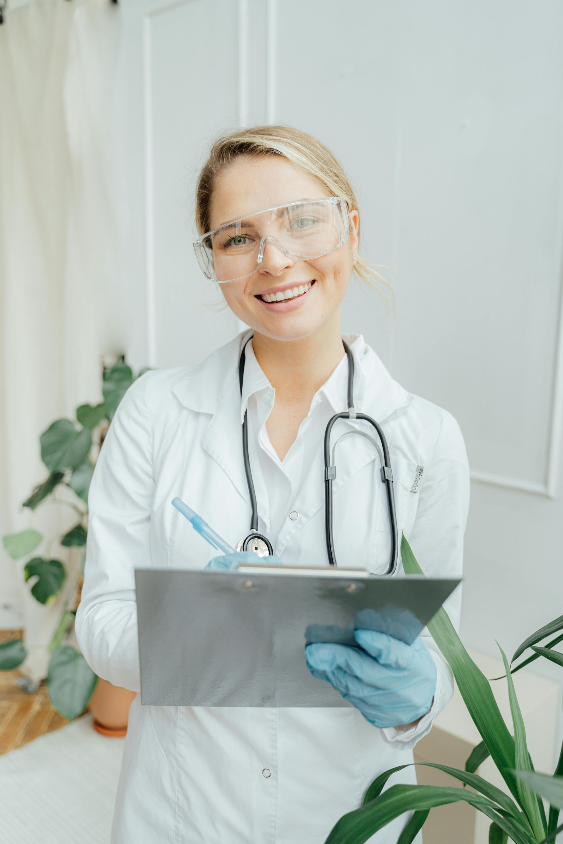 Smiling doctor with blonde hair, protective glasses, stethoscope, and clipboard, in a well-lit room with plants.