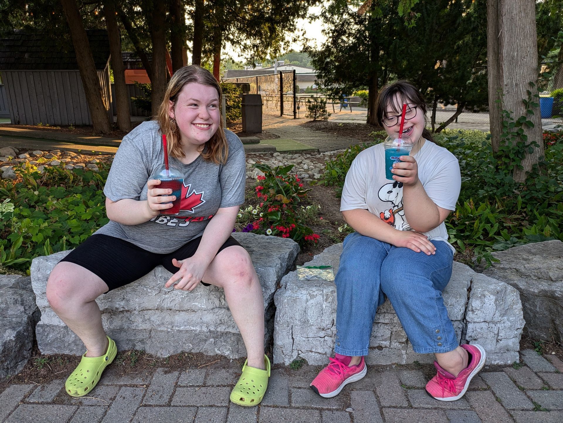 Two girls sitting on rocks, holding drinks with straws. They smile.