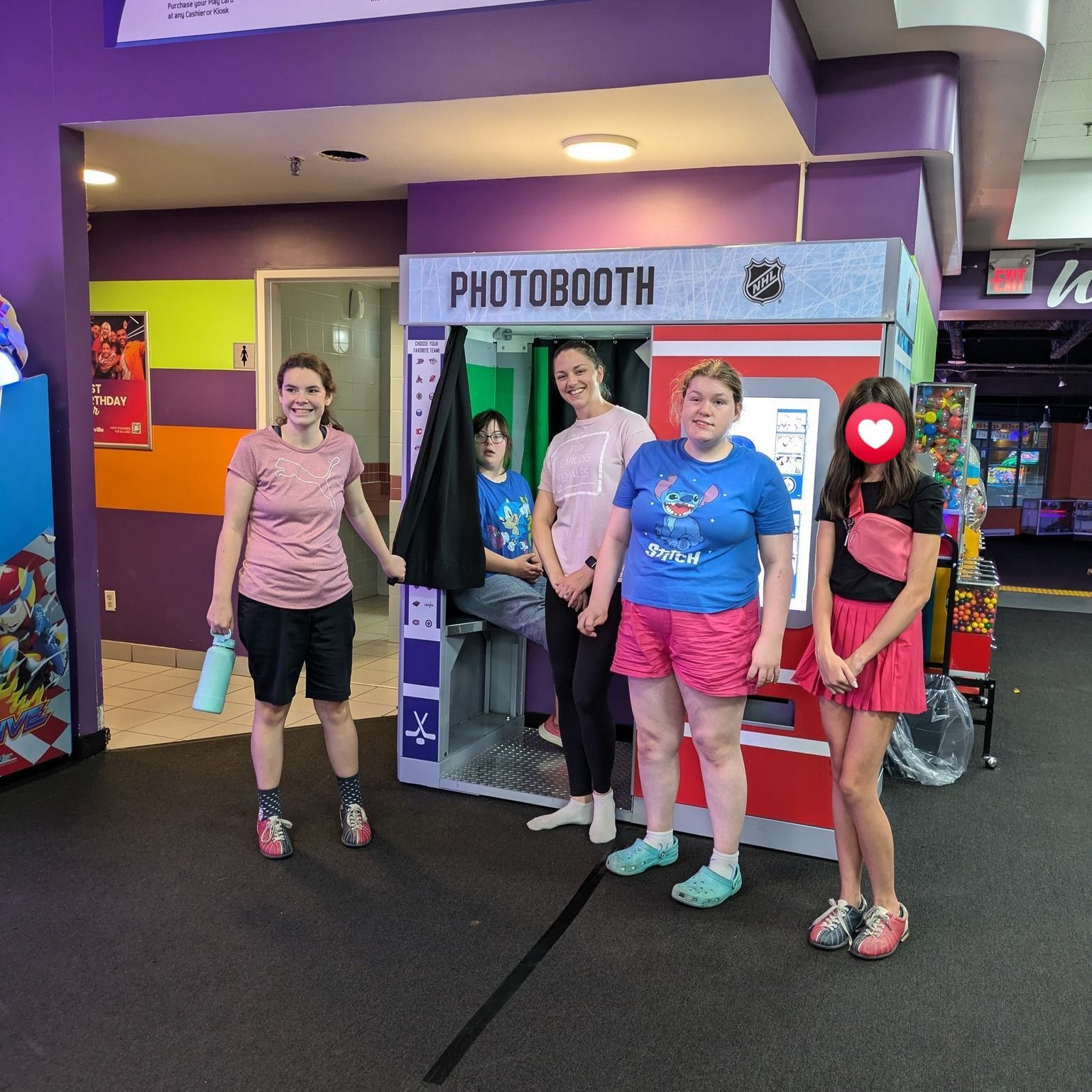 5 girls pose by a photobooth in an arcade. One person is in the booth, others outside smiling.