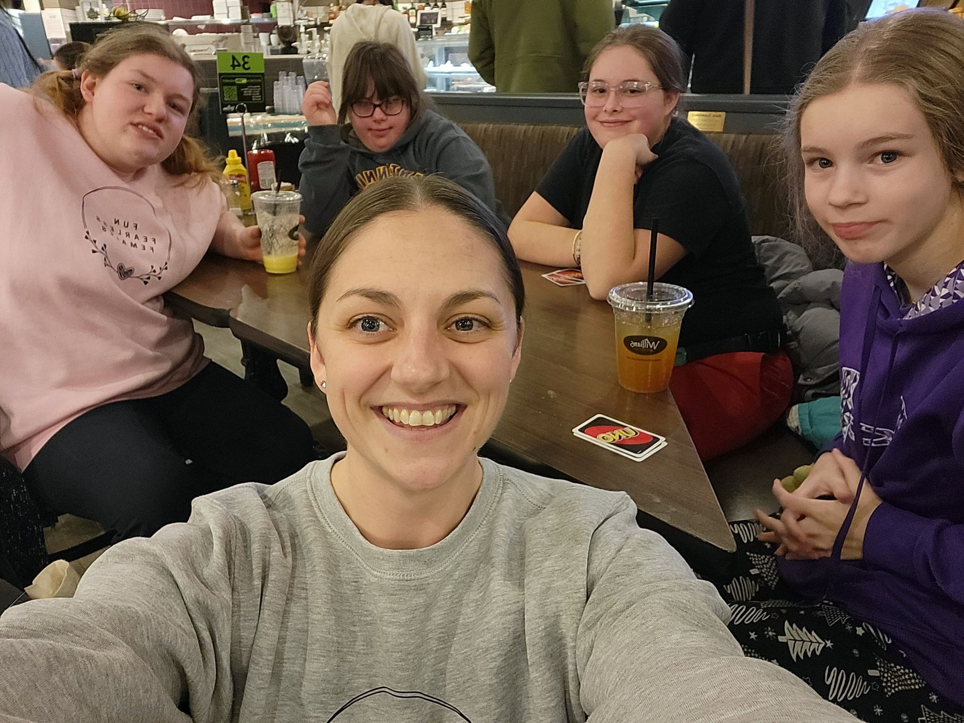Woman smiling, taking a selfie with four others at a table. Drinks, cards, and mustard on table.
