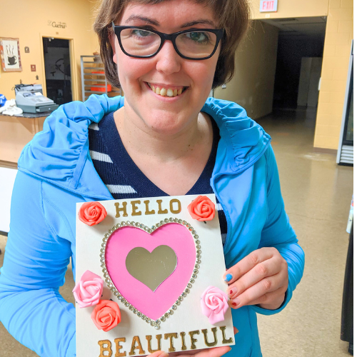Woman smiling, holding a framed craft with a pink heart cutout, “Hello Beautiful” text, and decorative roses.