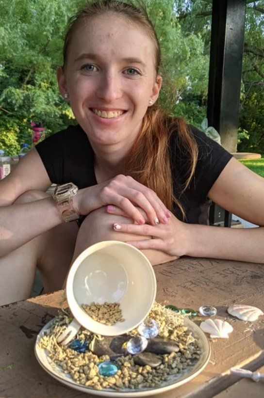 Woman smiling, looking at a small decorated plate with a tipped-over cup fairy garden; outdoors.