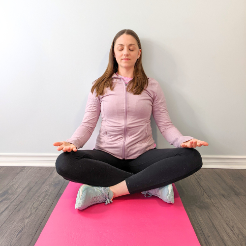 Woman in a seated meditation pose on a pink mat against a gray wall, hands open, eyes closed.