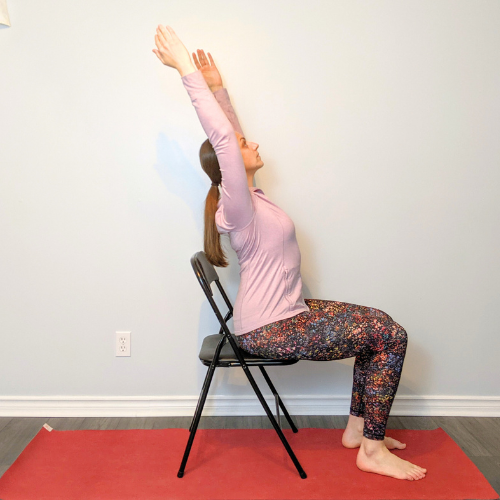 Woman in yoga pose, seated in a chair, arms extended upward. Wearing purple top and patterned pants.