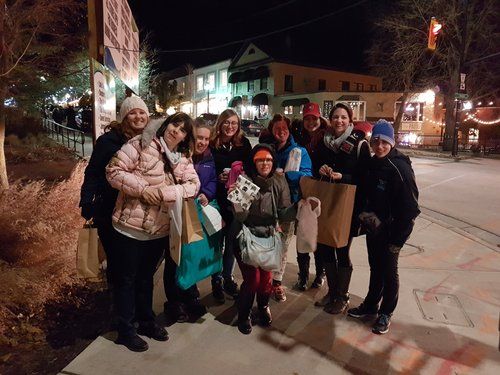 Group of women on a sidewalk at night, holding shopping bags, buildings and lights in background.