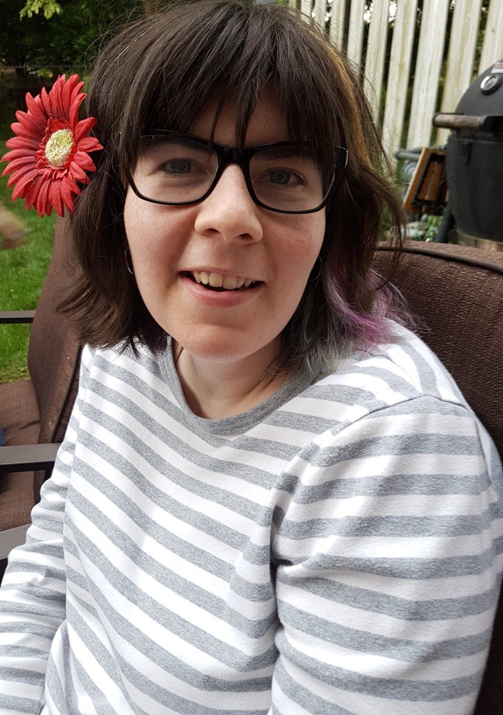 Woman with glasses, flower in hair, wearing striped shirt, smiling outdoors.