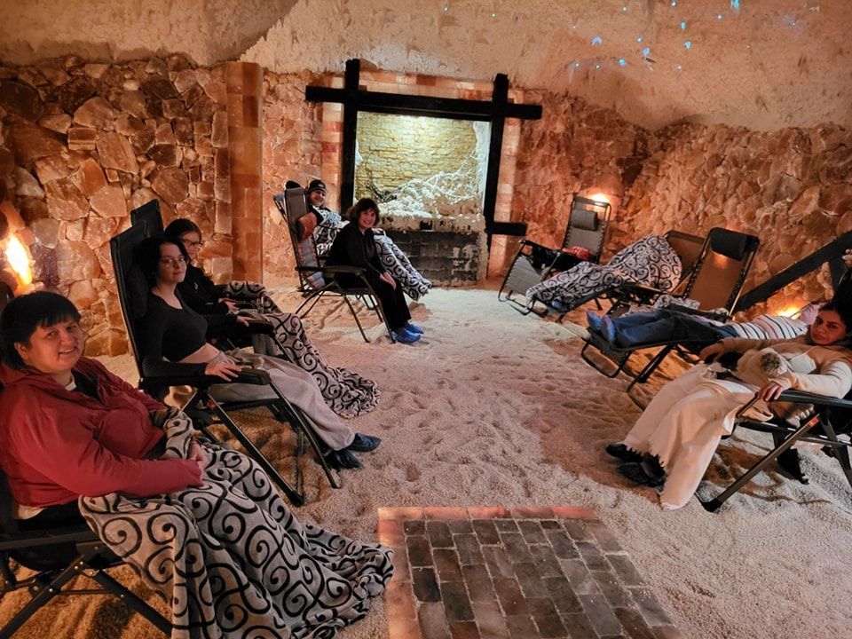 Women relaxing in a salt cave, seated in chairs, covered by blankets. The cave walls are salt.
