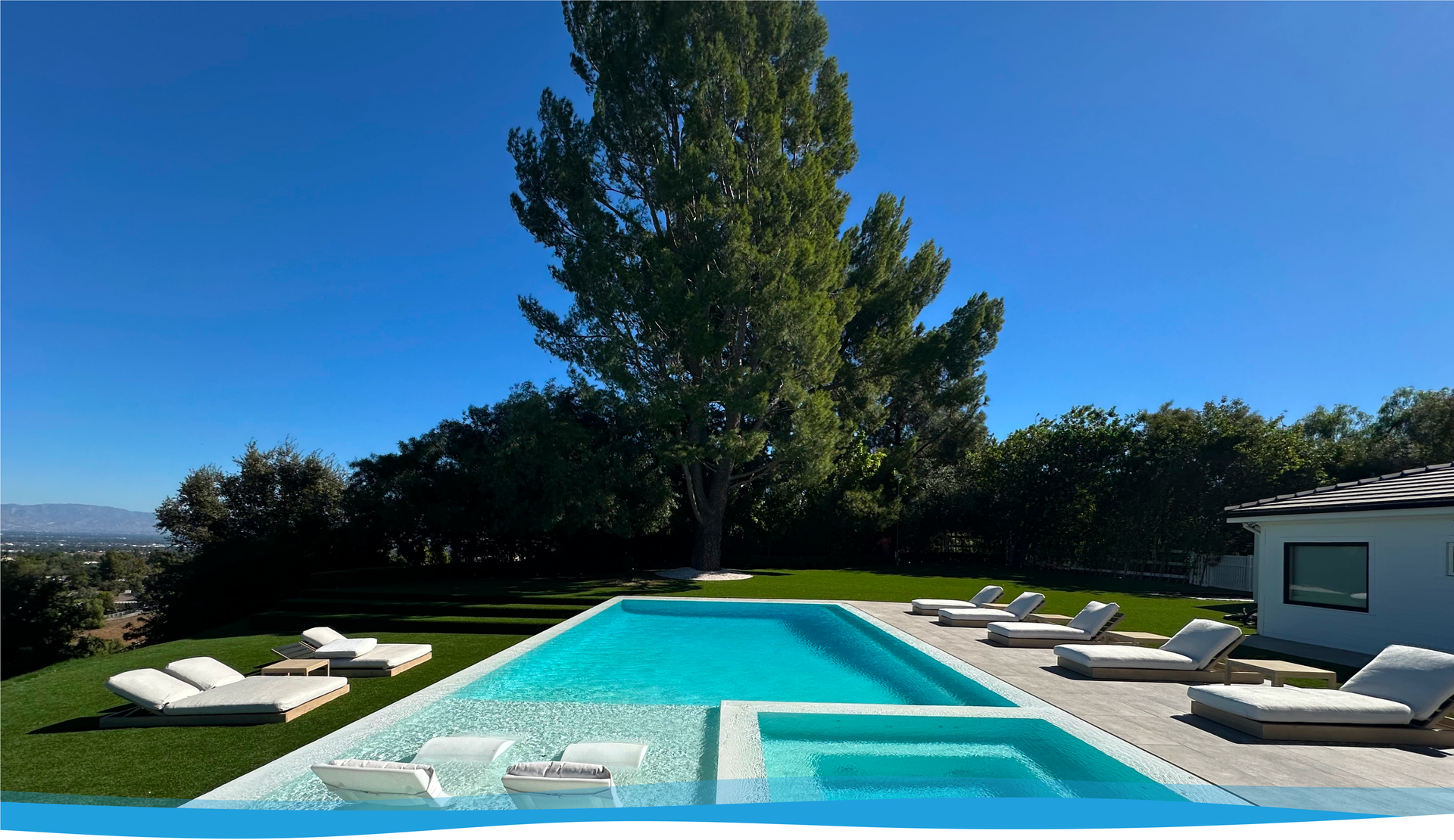 A rectangular swimming pool with lounge chairs and a tree on a sunny day.