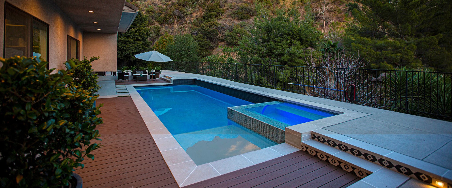 Outdoor swimming pool with spa and patio area, surrounded by lush greenery and trees.