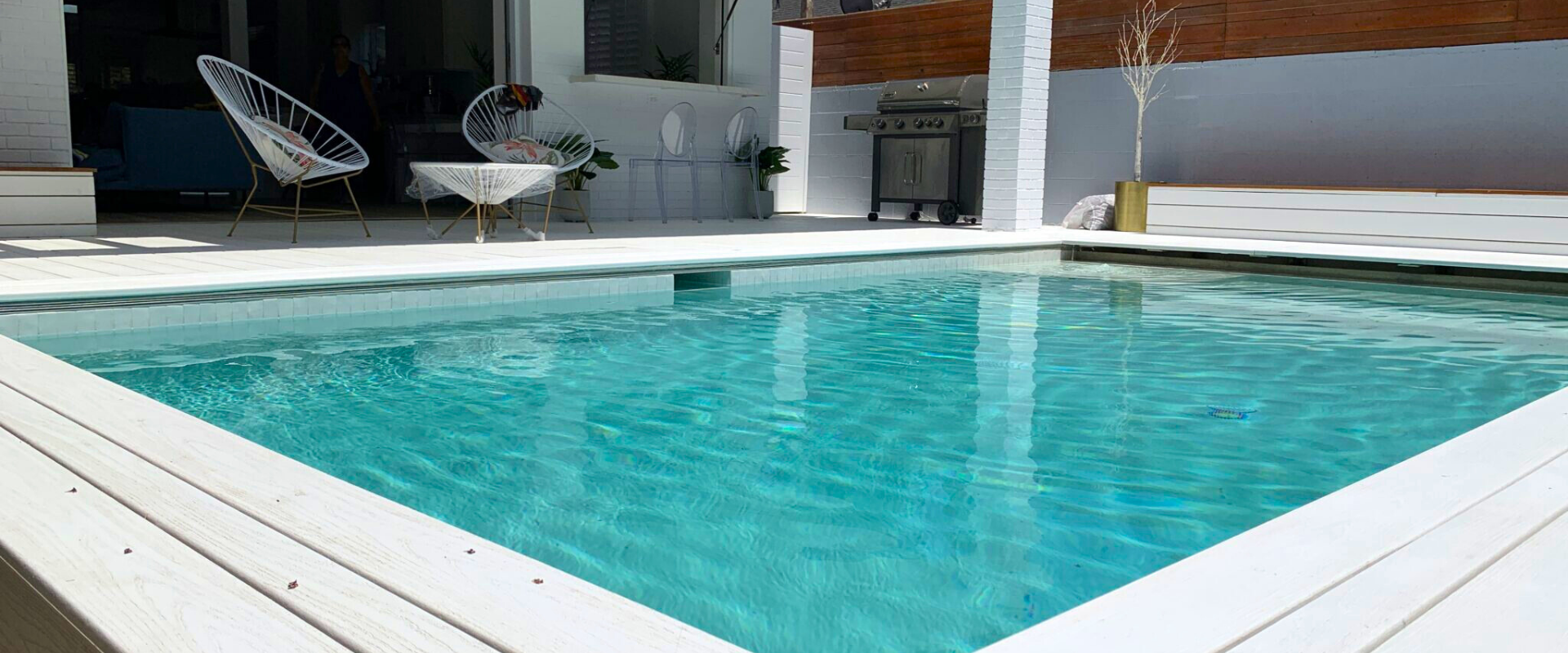 A turquoise pool with white decking in a backyard setting; a white table and chairs are in the background.