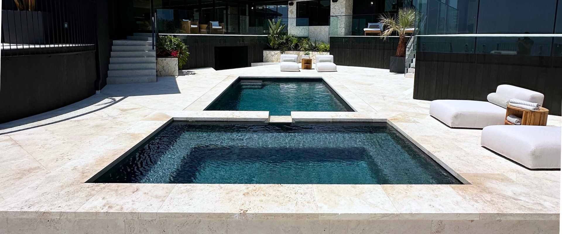 Two pools with dark water, surrounded by white stone patio. Lounge chairs near the pool, modern architecture.
