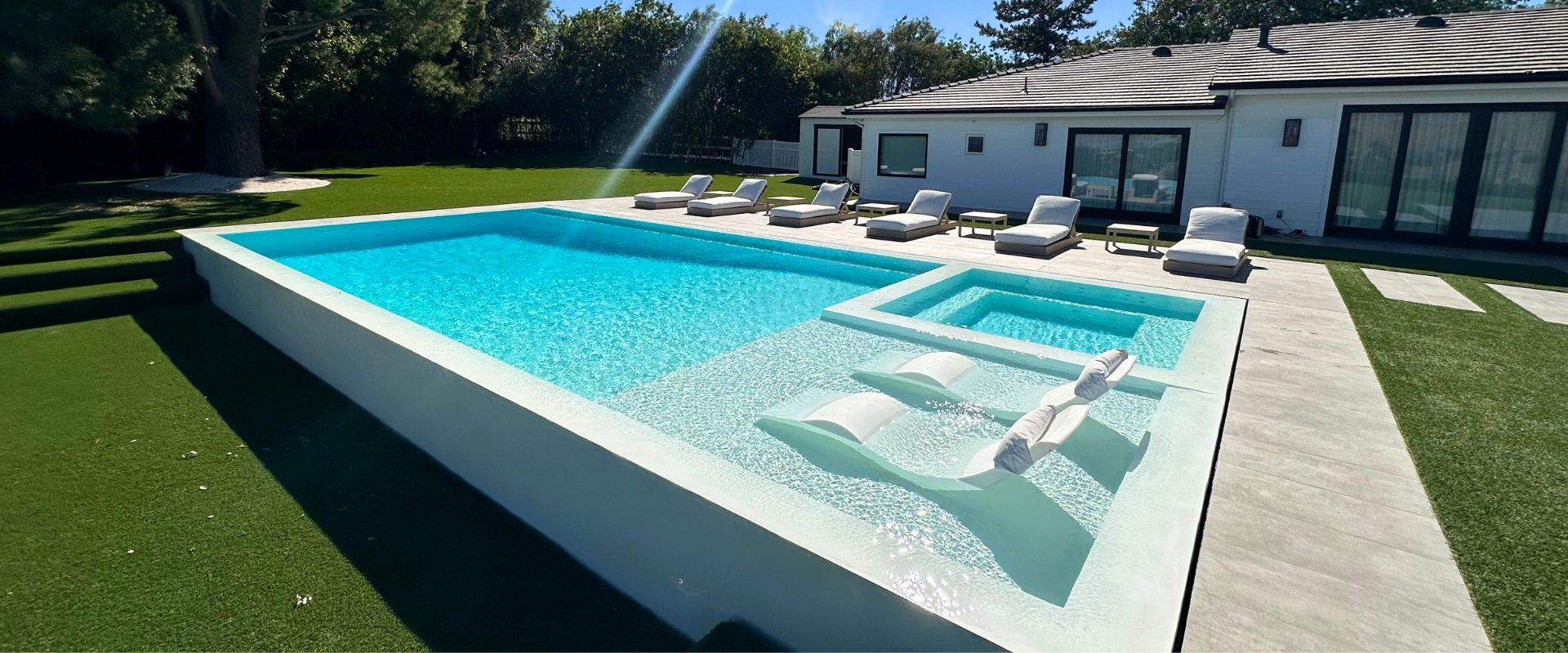 A modern pool with lounge chairs, a jacuzzi, and a house in the background. Lush green grass.