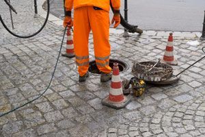 A Man In Orange Pants Is Standing Next To A Manhole Cover On A Cobblestone Street — NSL Plumbing and Gas in Banora Point, NSW
