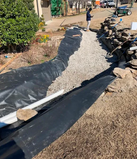 Gravel drainage trench lined with black fabric, with rocks stacked along one side and a person in the distance