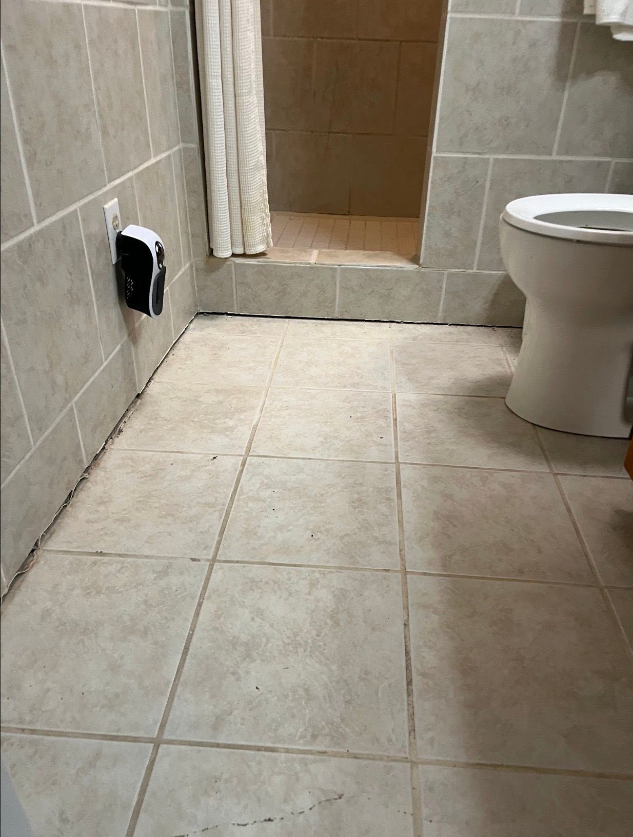 Small beige-tiled bathroom with a toilet, shower area, and wall vent opening.