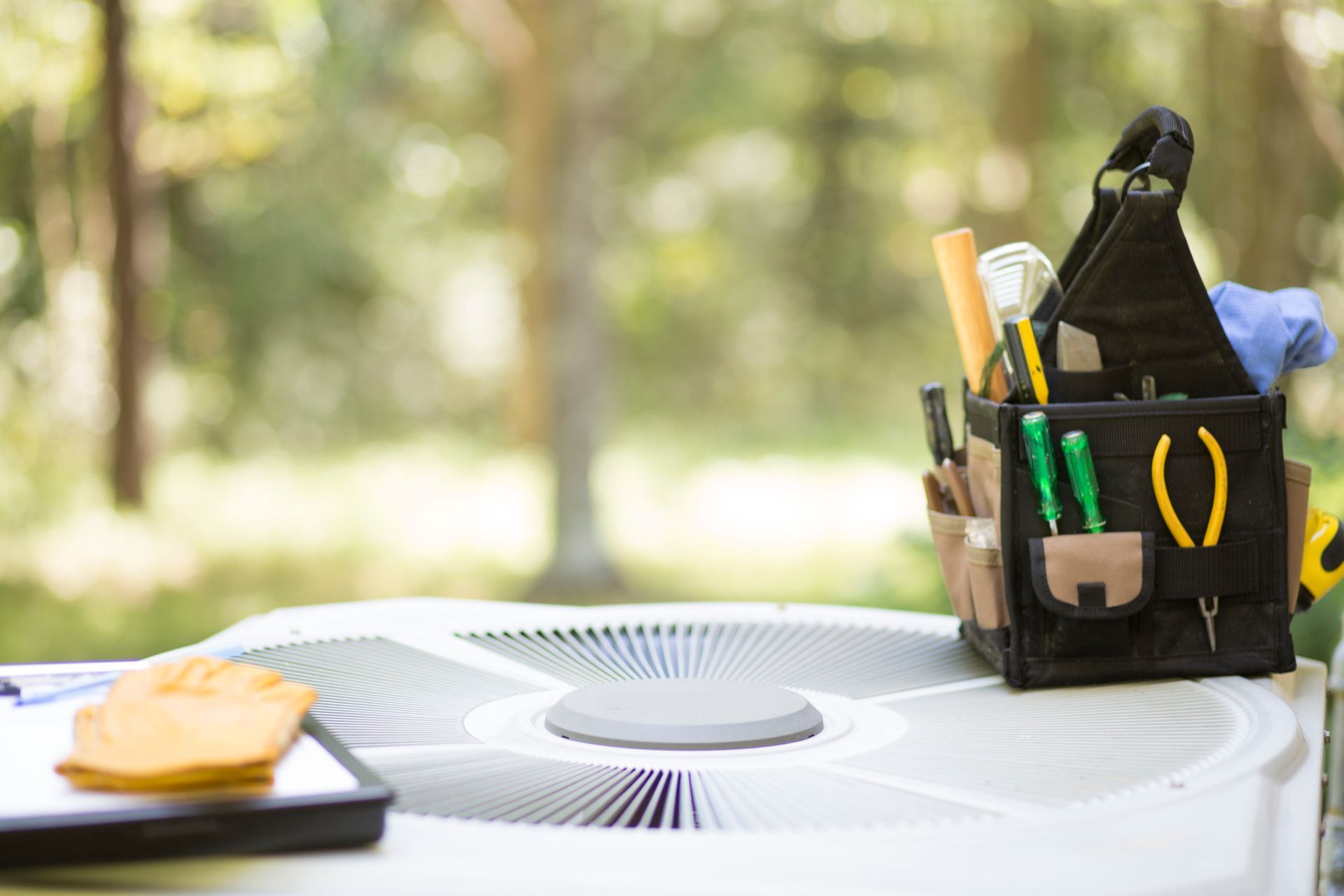 A tool bag and tablet with a worker’s glove sit on top of an outdoor AC unit.