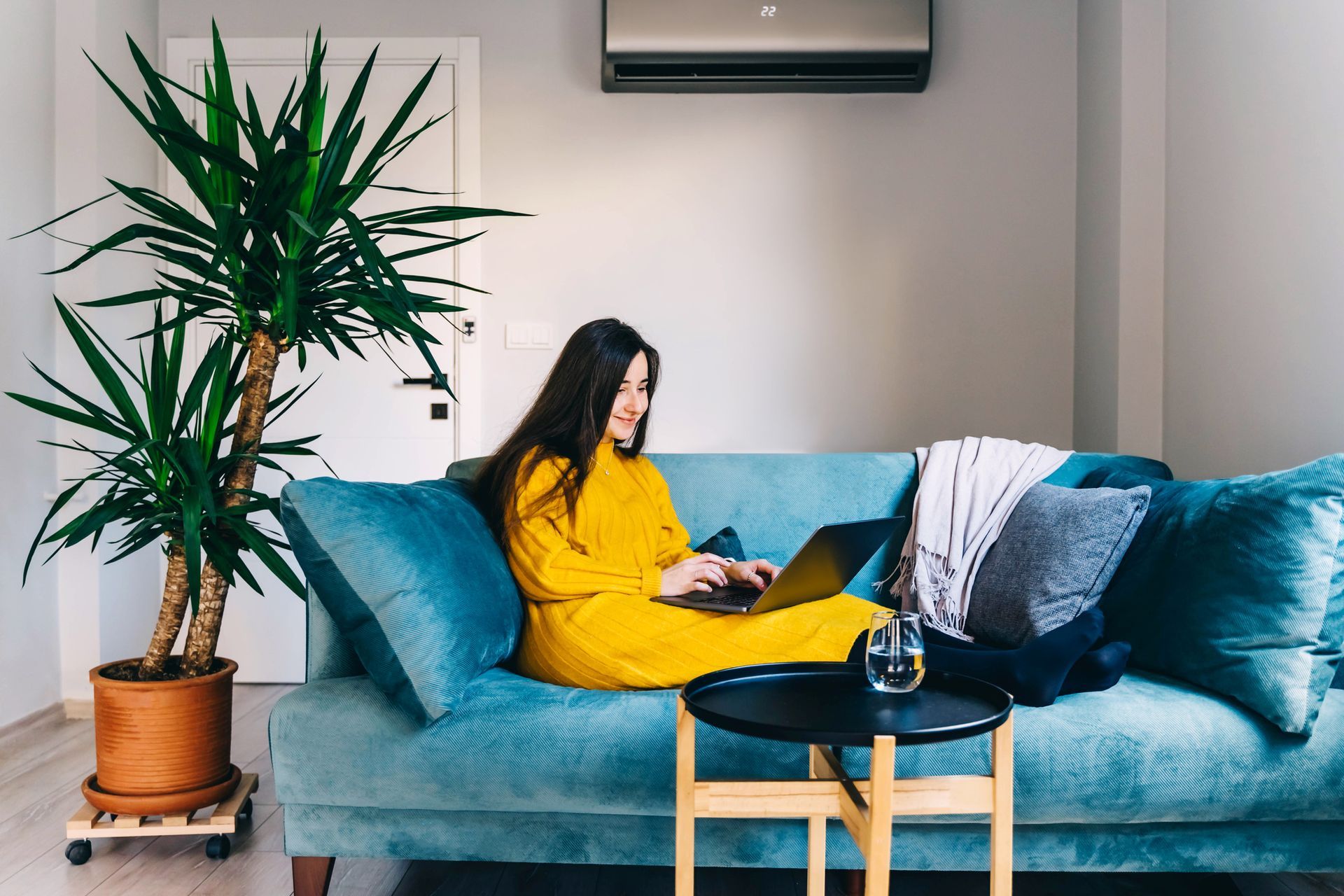 A woman sitting on a teal sofa using a laptop, with a potted plant and an air conditioner above.