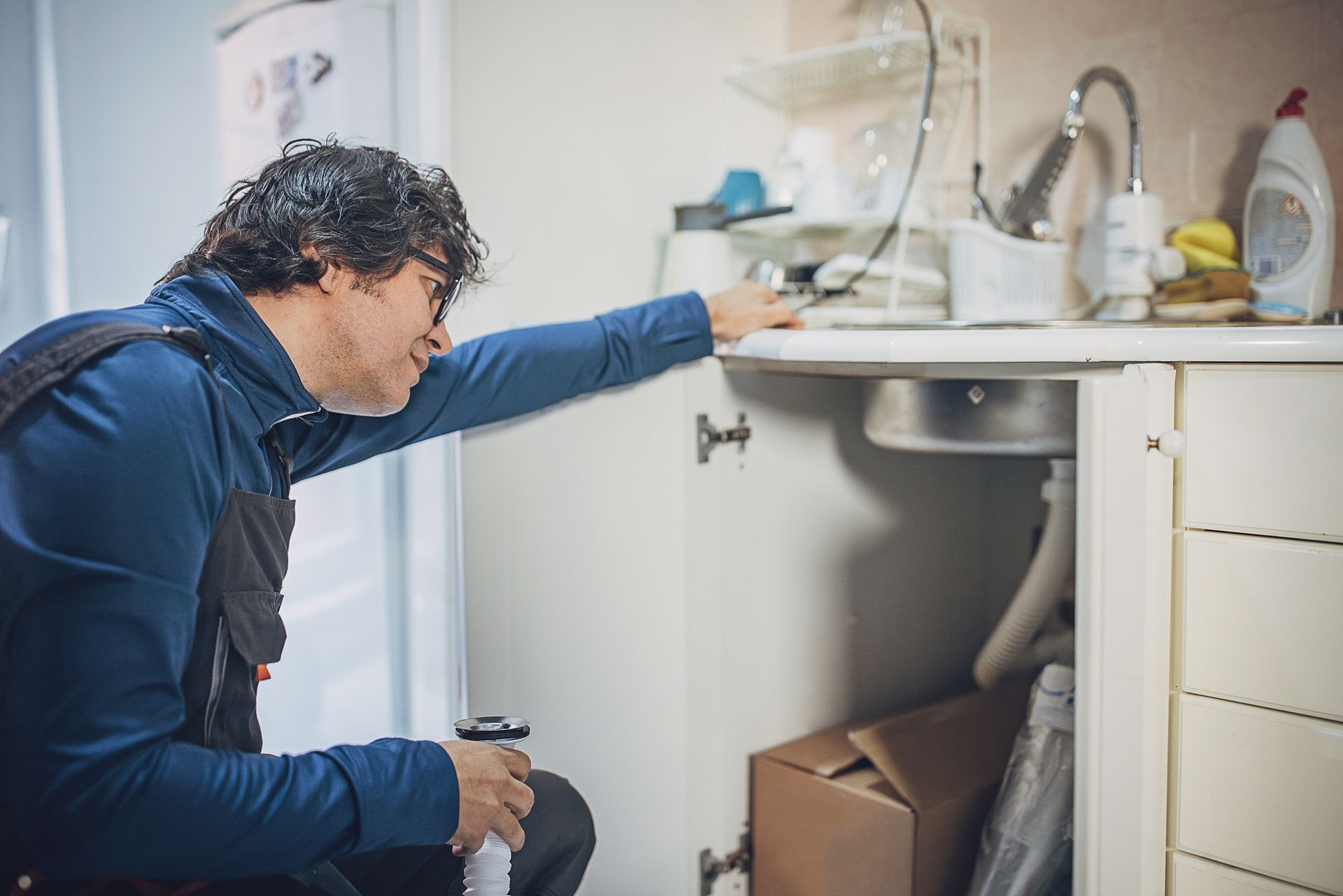 Man kneeling under a kitchen sink, looking at plumbing. He wears blue and glasses, and holds a pipe.
