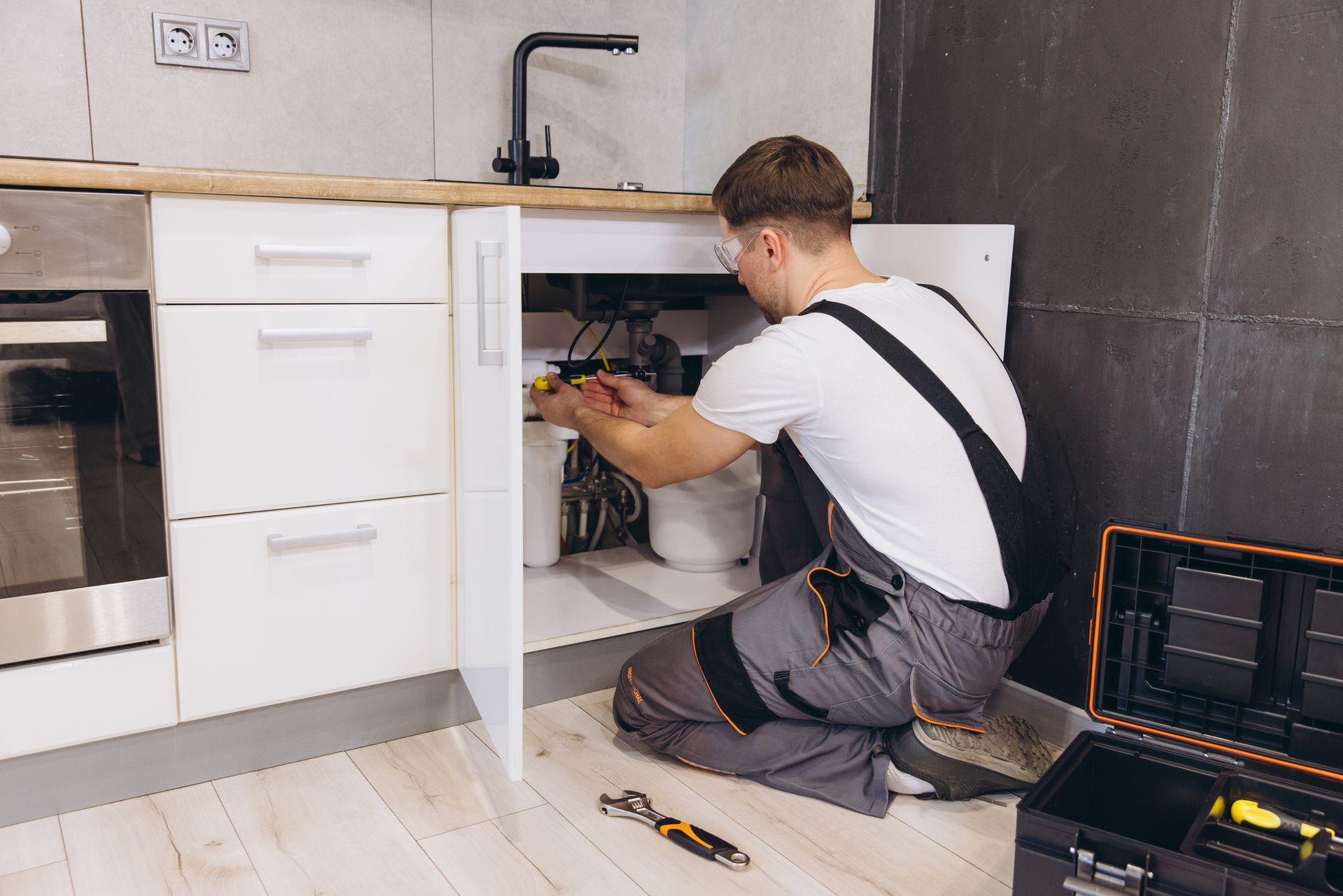 A man in work overalls is working on plumbing under a kitchen sink.