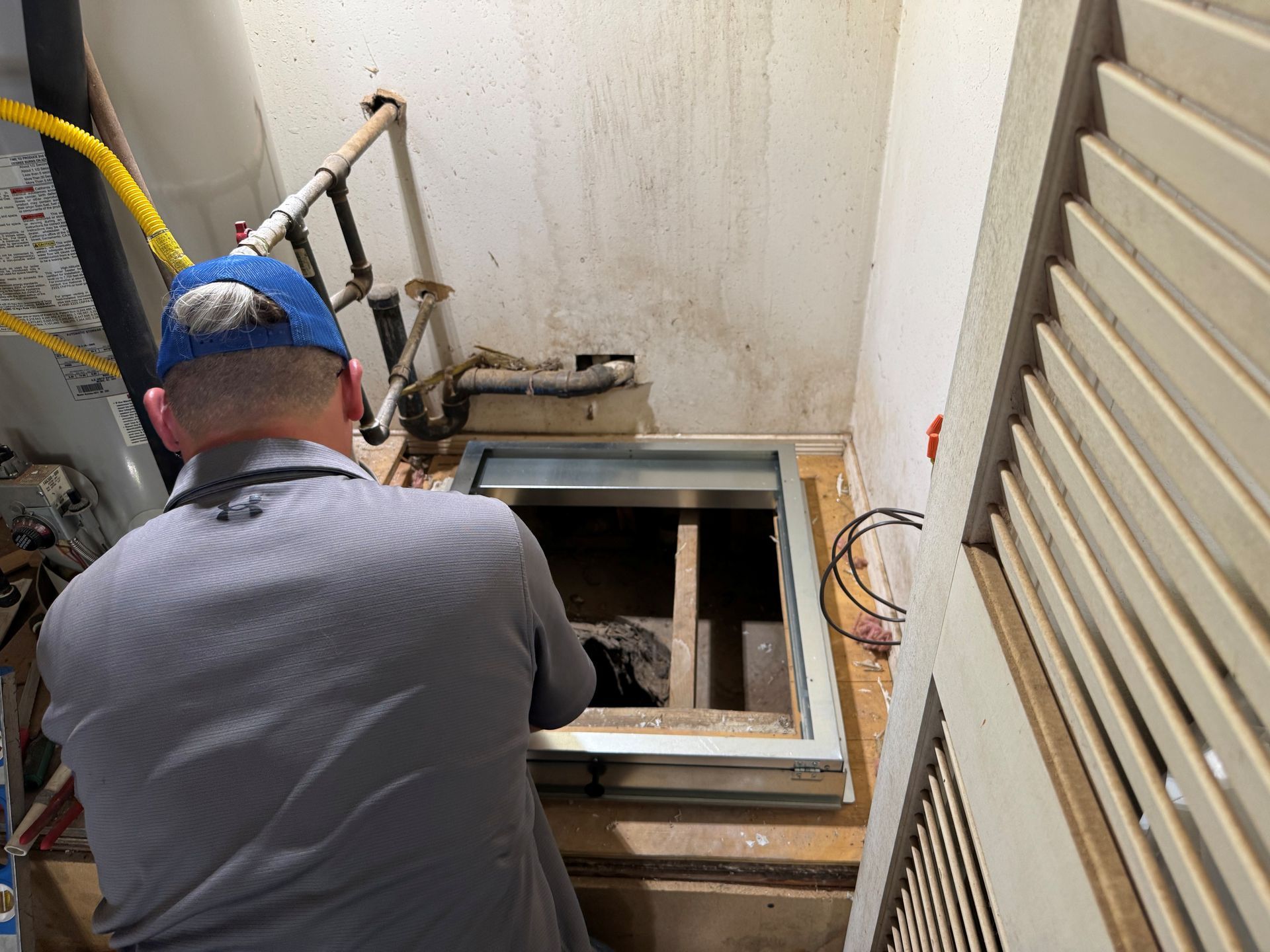 Man in blue hat looks into open hatch in a utility room. Yellow tube and pipes are visible.