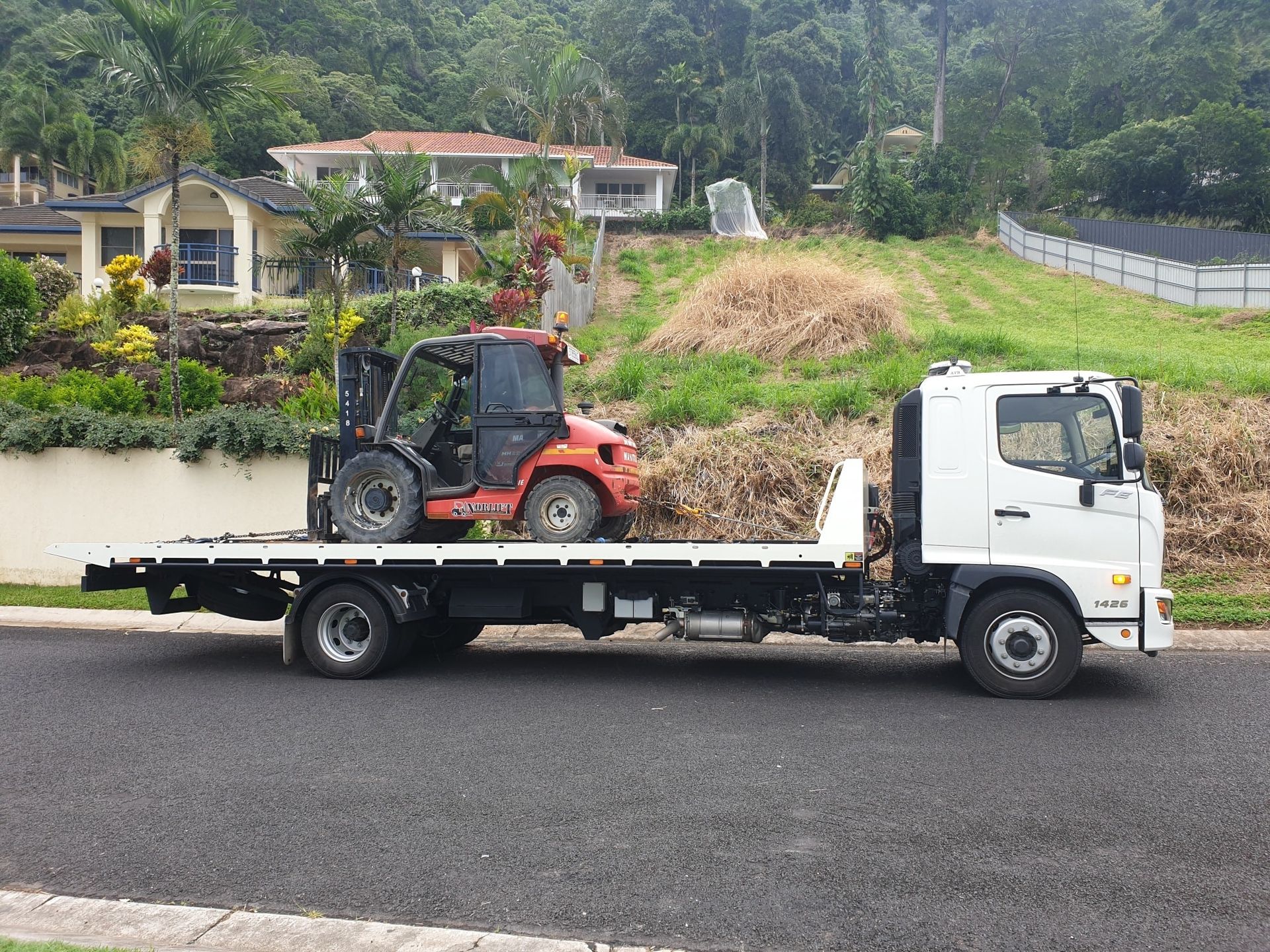 a Flatbed Truck Carrying an Orange Forklift on a Residential Street — Tigo’s Towing In Cairns City, QLD