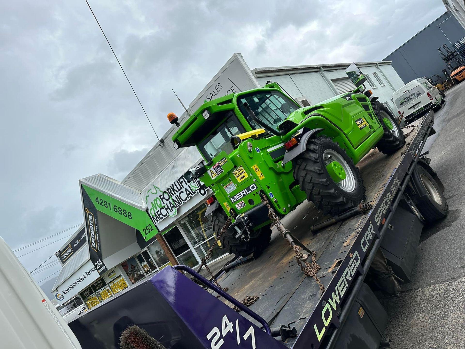 Green Merlo Telehandler on a Flatbed Tow Truck. Shop in Background — Tigo’s Towing In Cairns City, QLD
