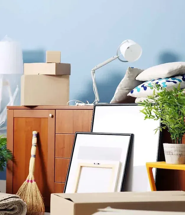 A living room filled with boxes , frames , pillows , a lamp and a broom.