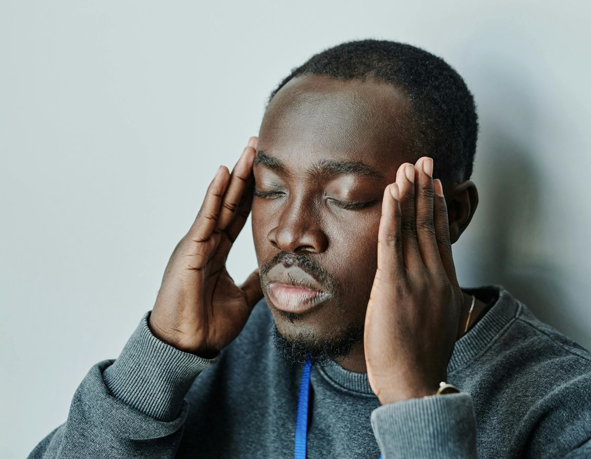 Man with dark skin, eyes closed, pressing temples, wearing gray sweater.