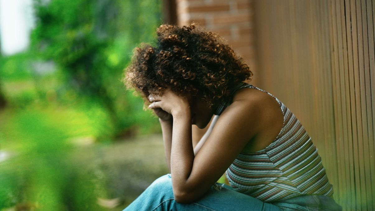 Woman with curly hair, sitting, head in hands, looking down; expression suggests sadness.