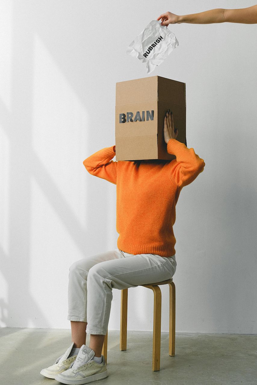 A woman is sitting on a stool with a cardboard box on her head.