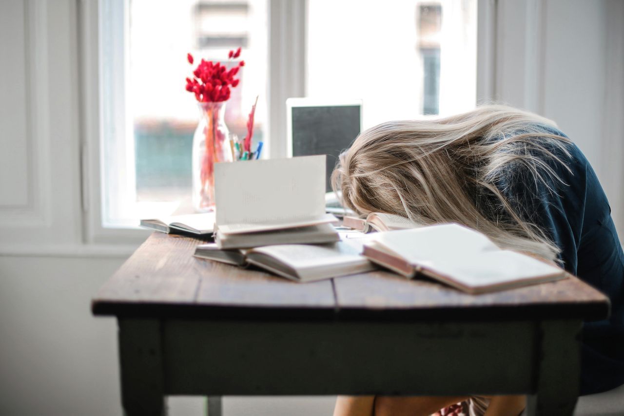 Woman with blonde hair resting her head on a desk cluttered with books and a laptop, near a window.