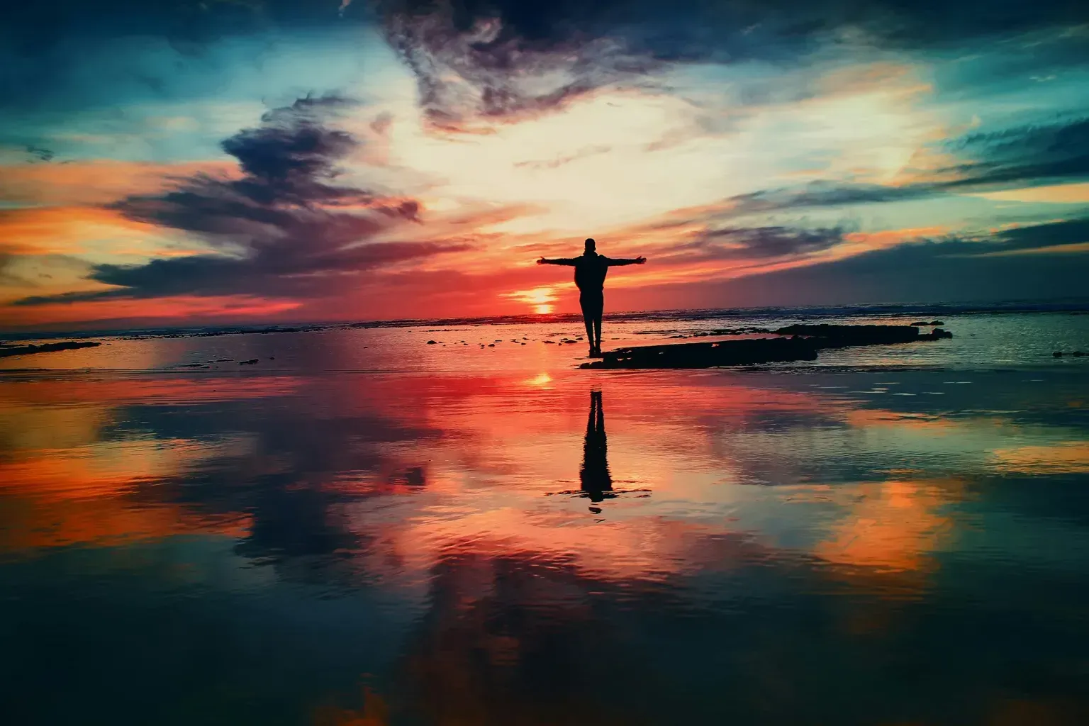 A silhouette of a person standing on a beach at sunset with their arms outstretched.
