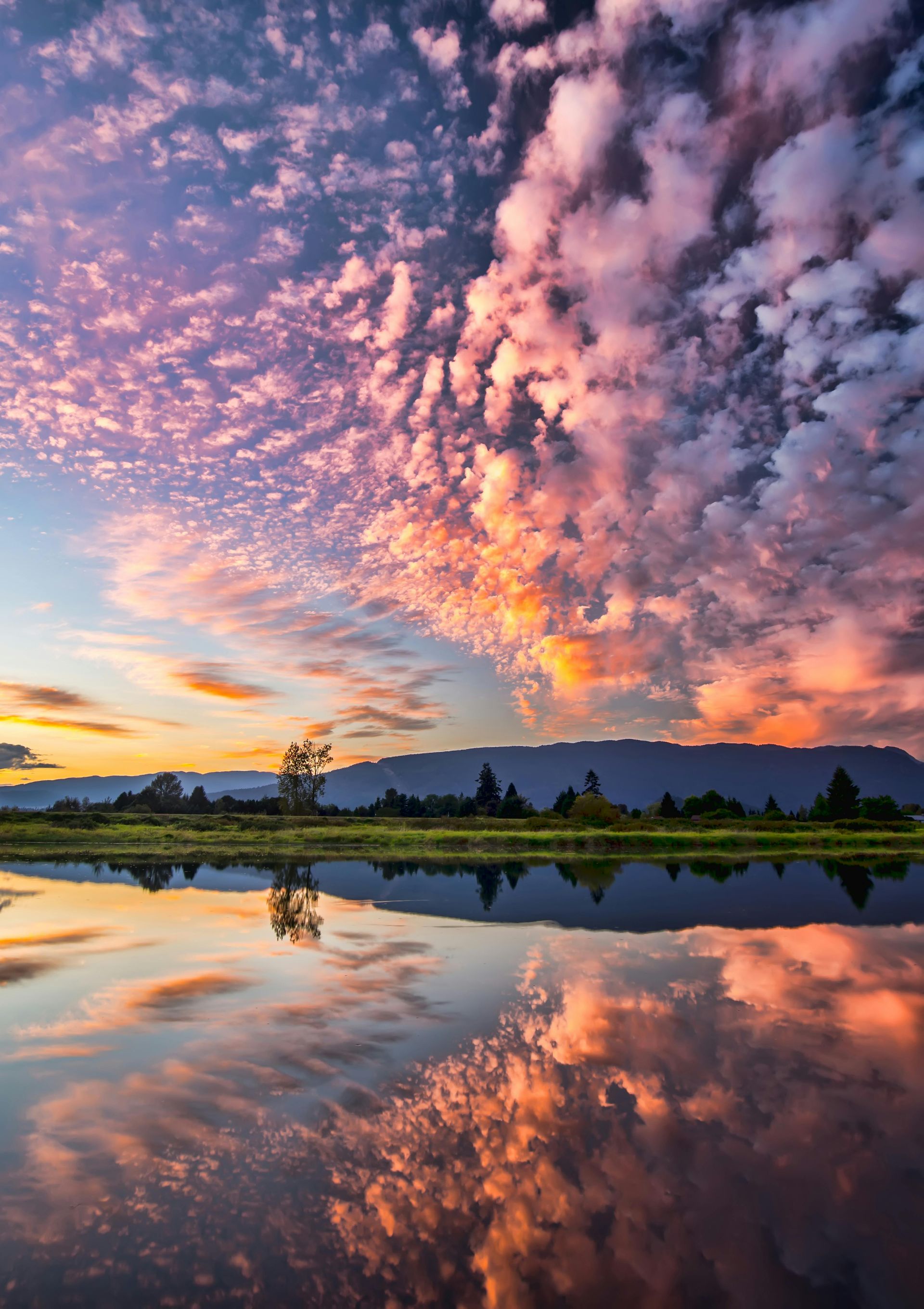 A sunset over a lake with a mountain in the background and clouds reflected in the water.