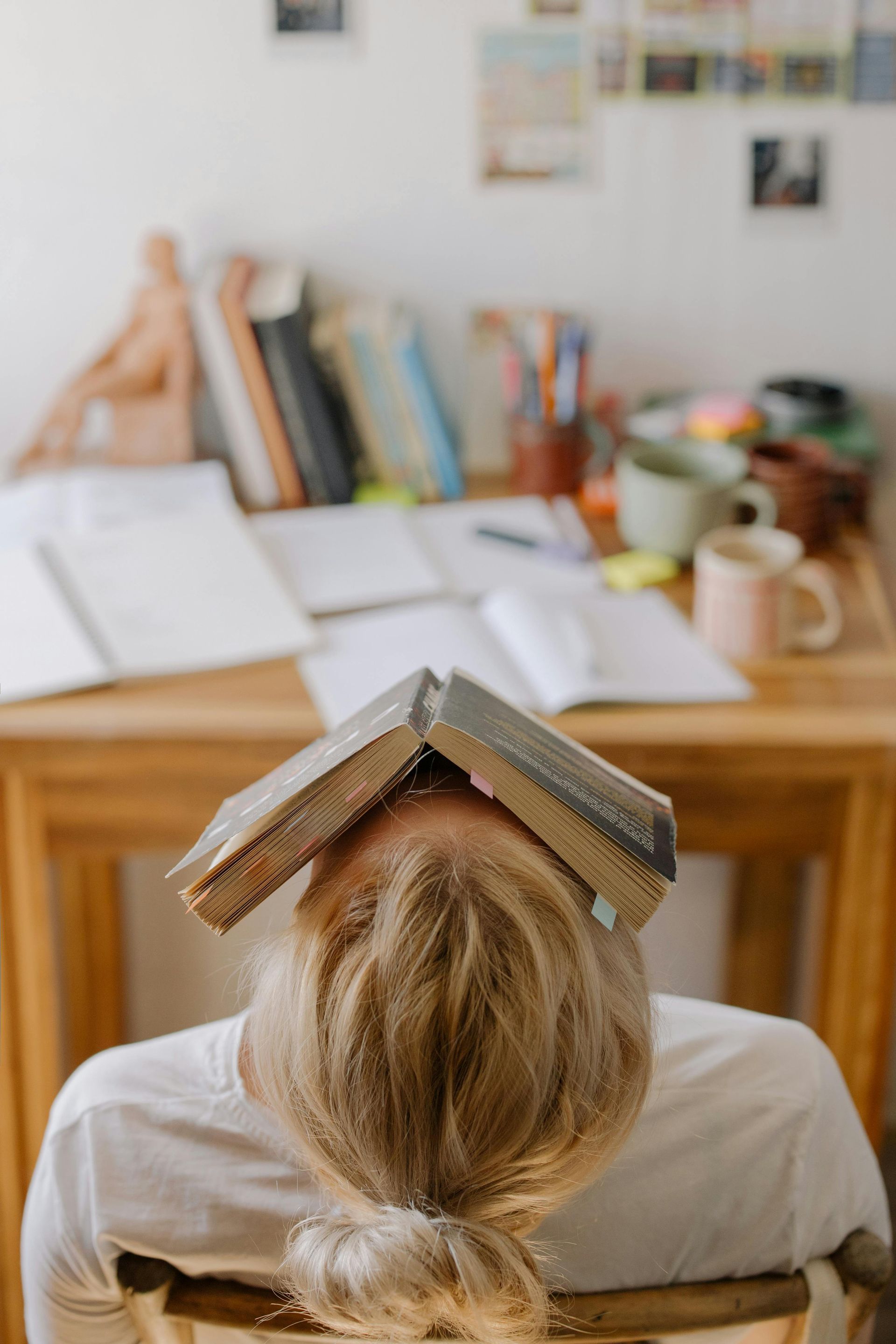 Person with a book on their head, slumped at a cluttered desk. Blonde hair, white shirt, wooden table.