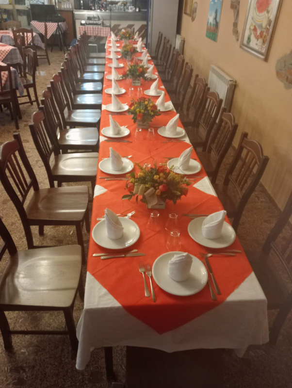 Lungo tavolo da pranzo apparecchiato per un pasto, tovaglia arancione, piatti e tovaglioli bianchi, mazzi di fiori.