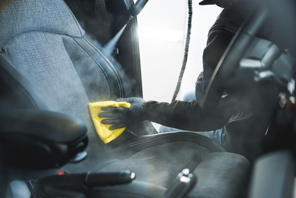 Persona che pulisce a vapore il sedile di un'auto con un panno giallo, vapore visibile, interni neri.