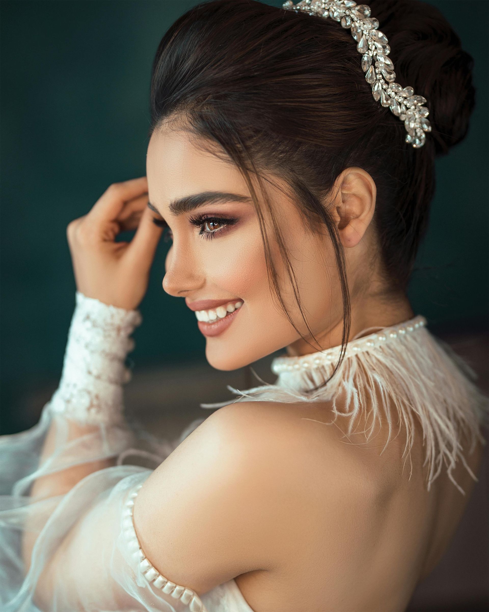 Donna sorridente con elegante acconciatura raccolta, che indossa un abito bianco con dettagli di piume e perle.