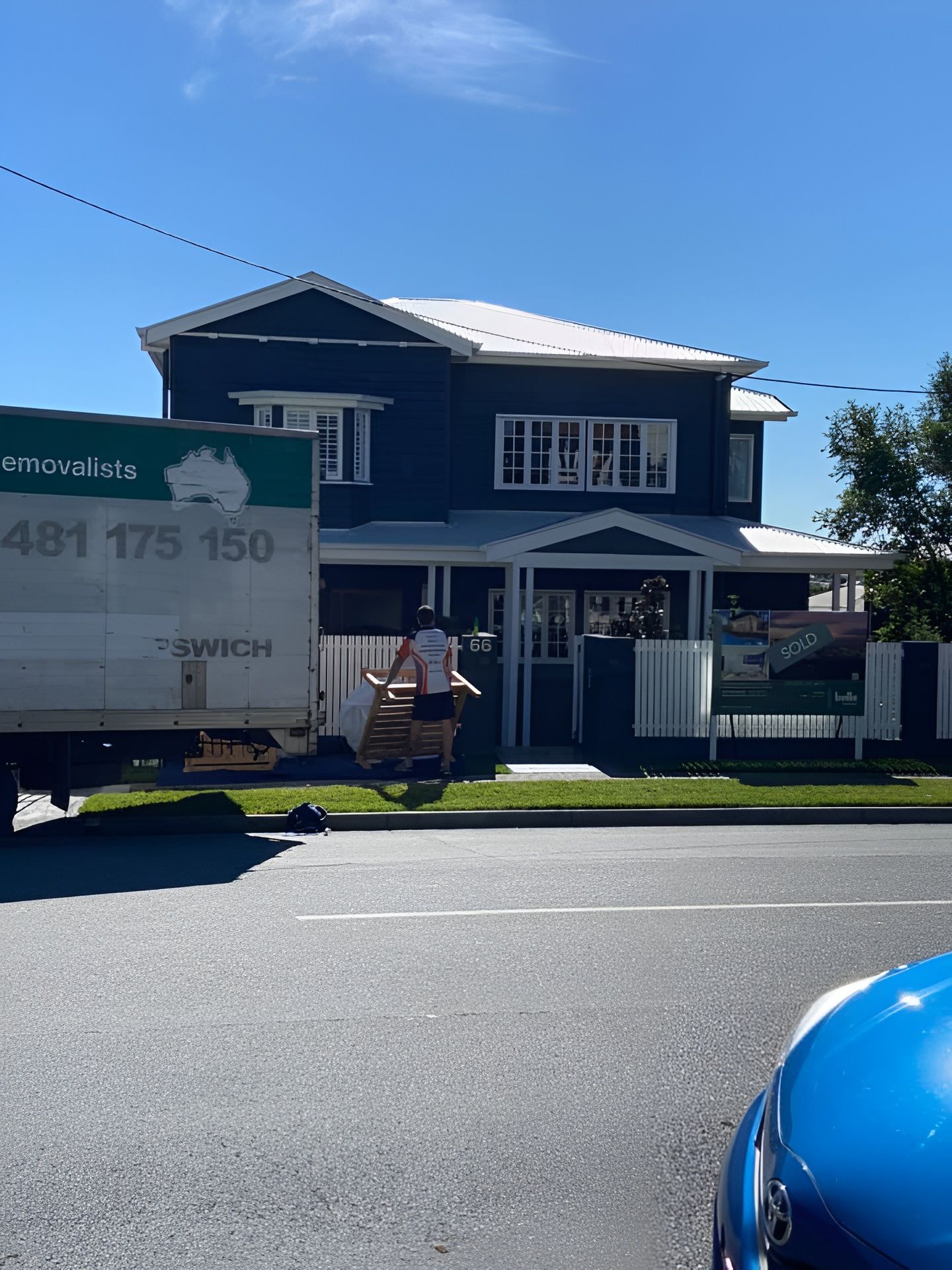 Person carrying items towards a two-story blue house, moving truck parked on the street. — Gantic Removals Ipswich in Ipswich, QLD
