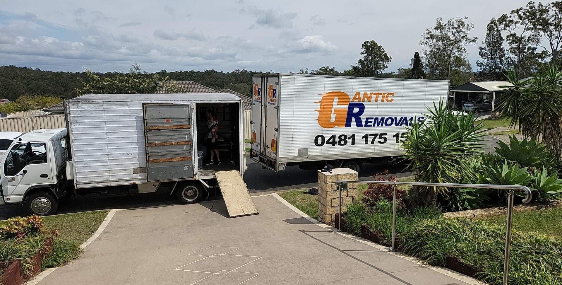 Two moving trucks parked in driveway, ramp down. One truck is white, the other has blue logo
