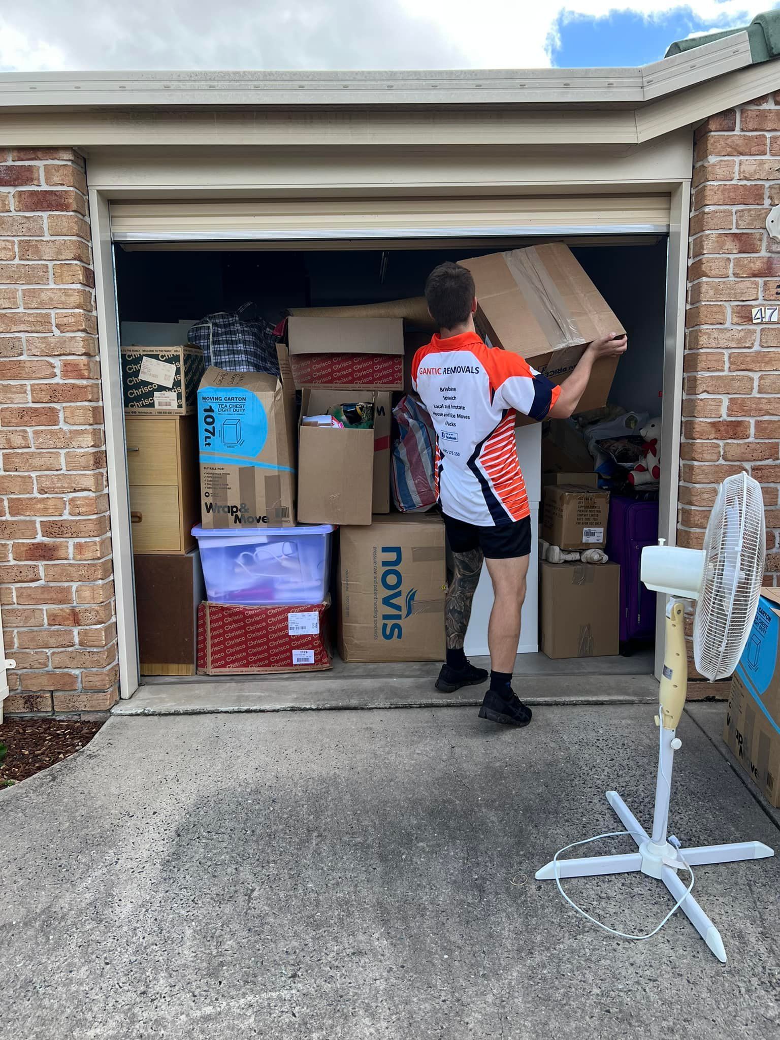 A person in athletic gear loads a cardboard box into a garage filled with packed boxes and storage bins. — Gantic Removals Ipswich in Ipswich, QLD