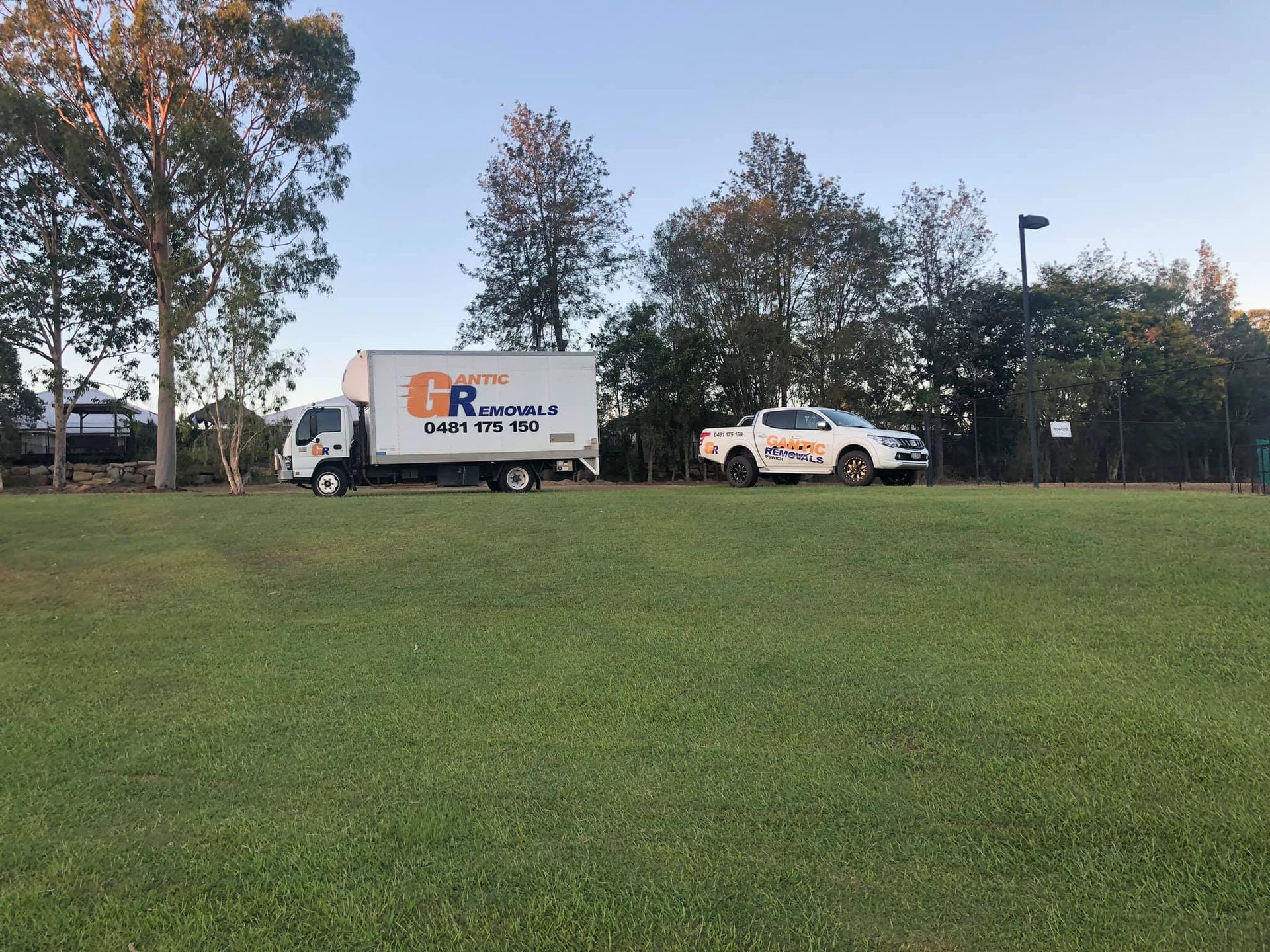 White moving truck and a white pickup truck on a grassy area, trees in the background under a blue sky. — Gantic Removals Ipswich in Ipswich, QLD