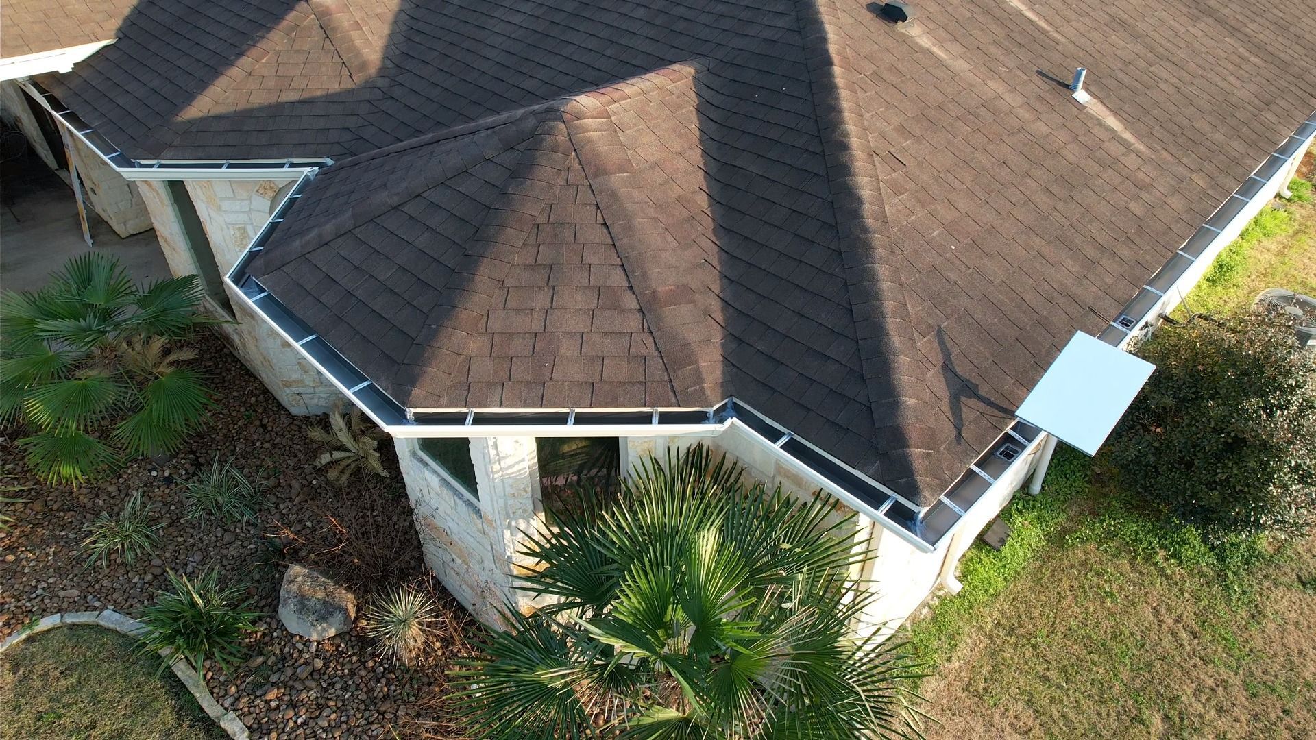 Overhead view of a house with a brown shingled roof, light stone walls, and surrounding greenery.