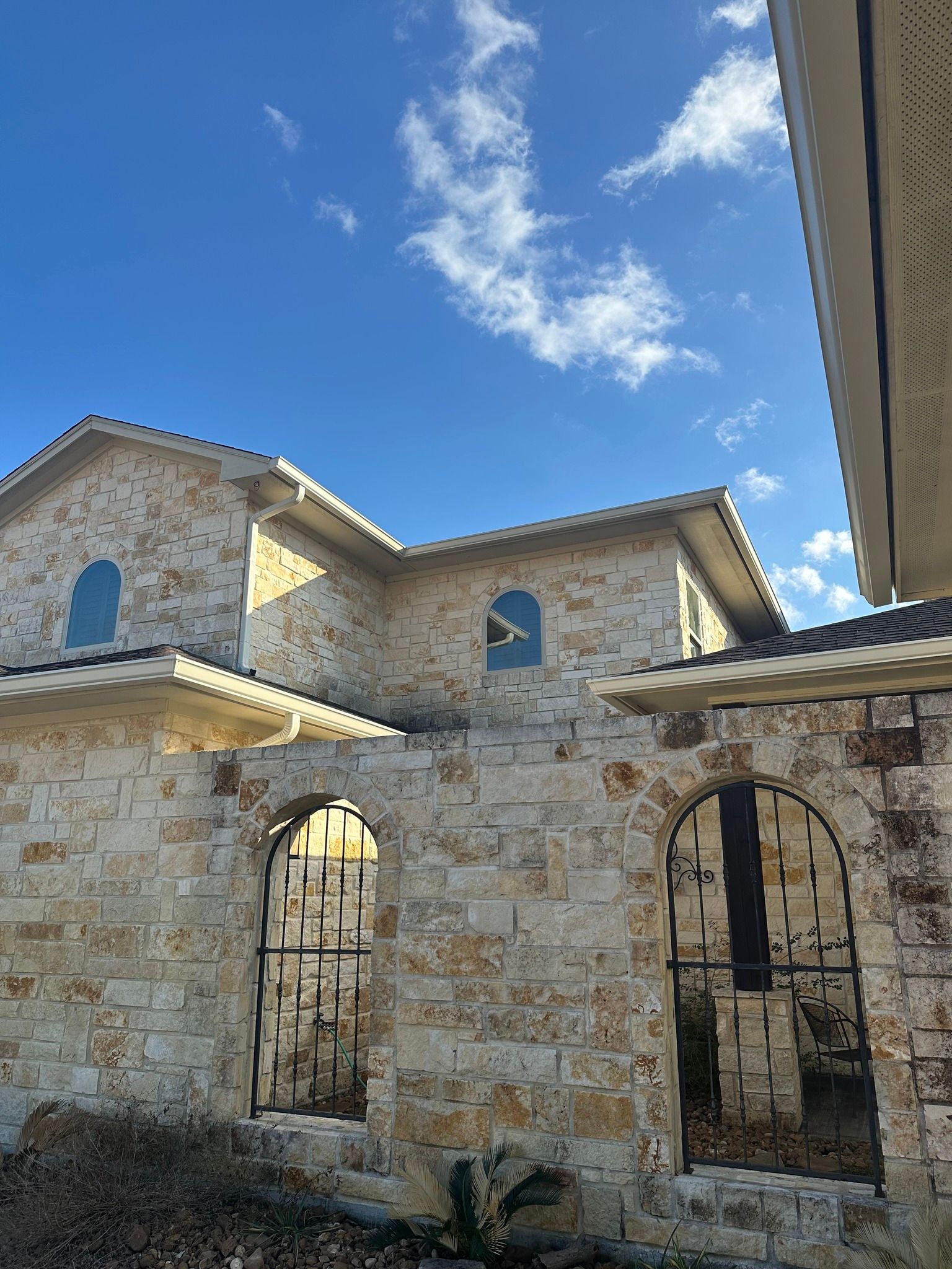 Stone house with arched windows and black metal gates under a blue sky with clouds.