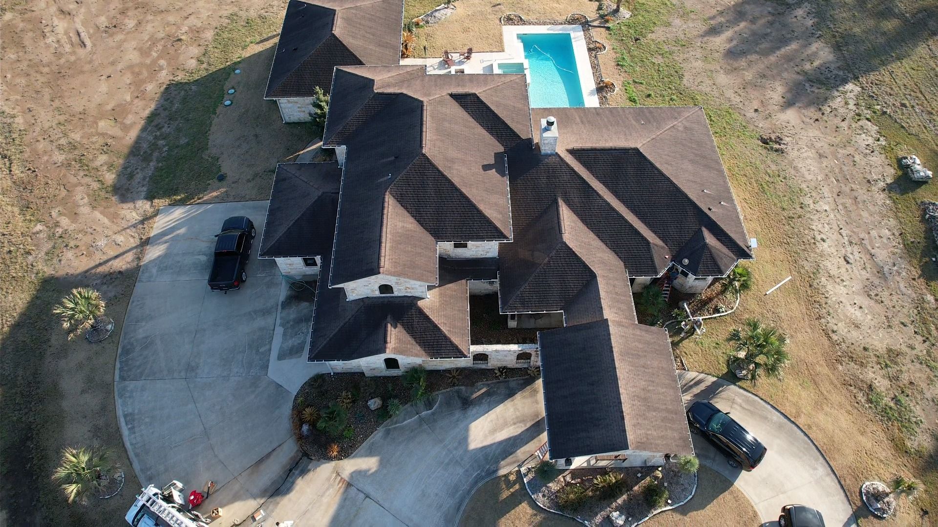 Aerial view of a large, brown-roofed house with a pool, vehicles, and a circular driveway on a dry landscape.