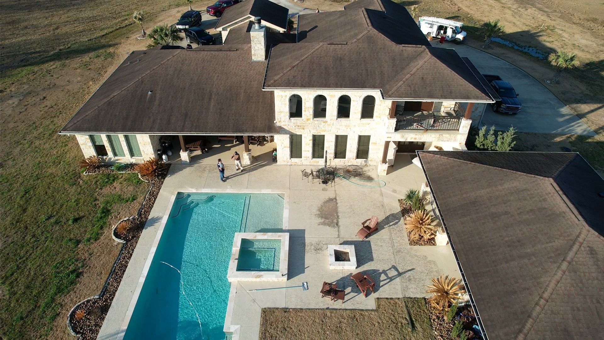 Aerial view of a large house with a pool and patio. Brown roof, beige stone exterior. Cars parked in the driveway.