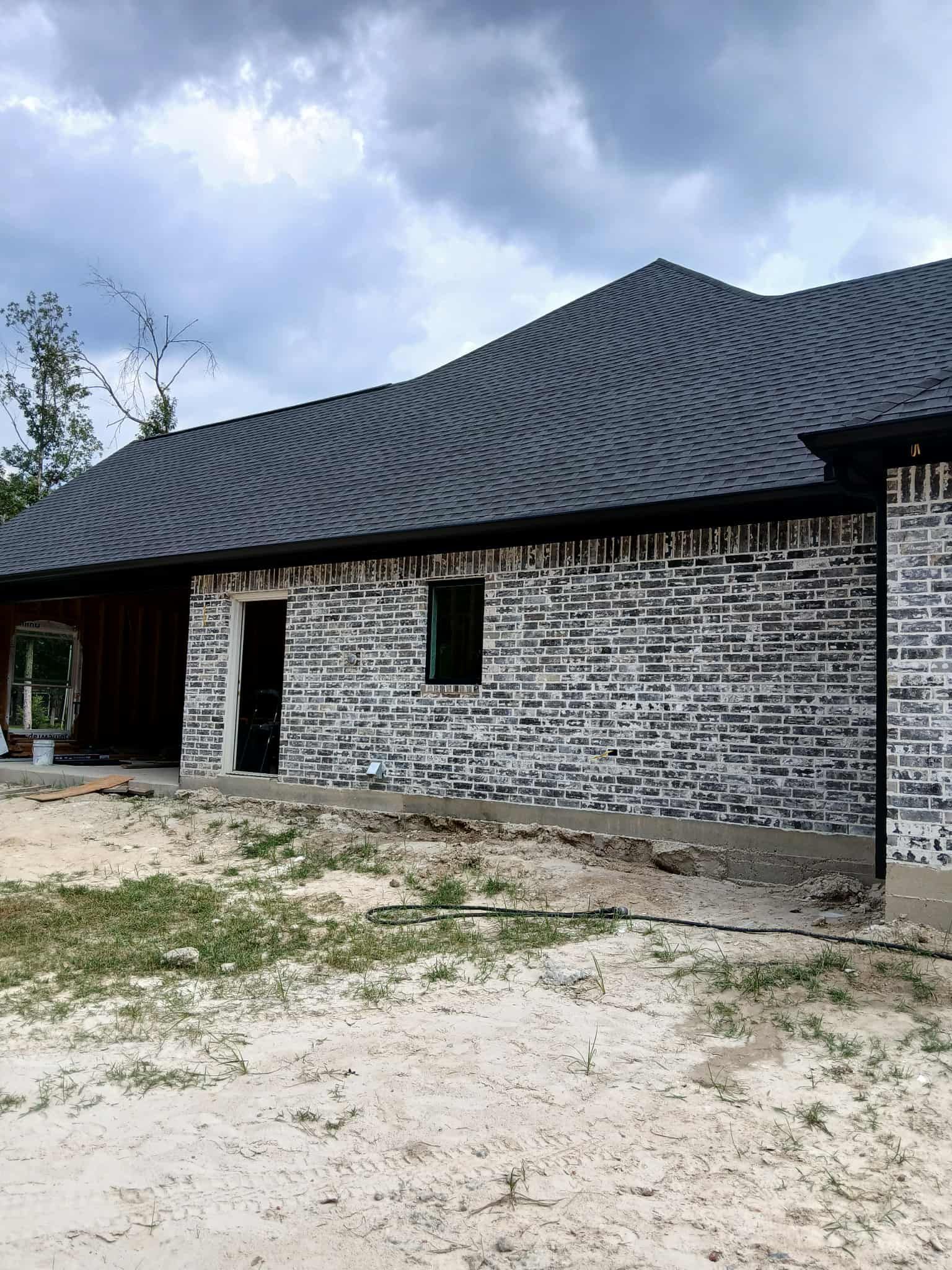 New construction home with dark brick walls, gray roof, and sandy ground. Overcast sky.