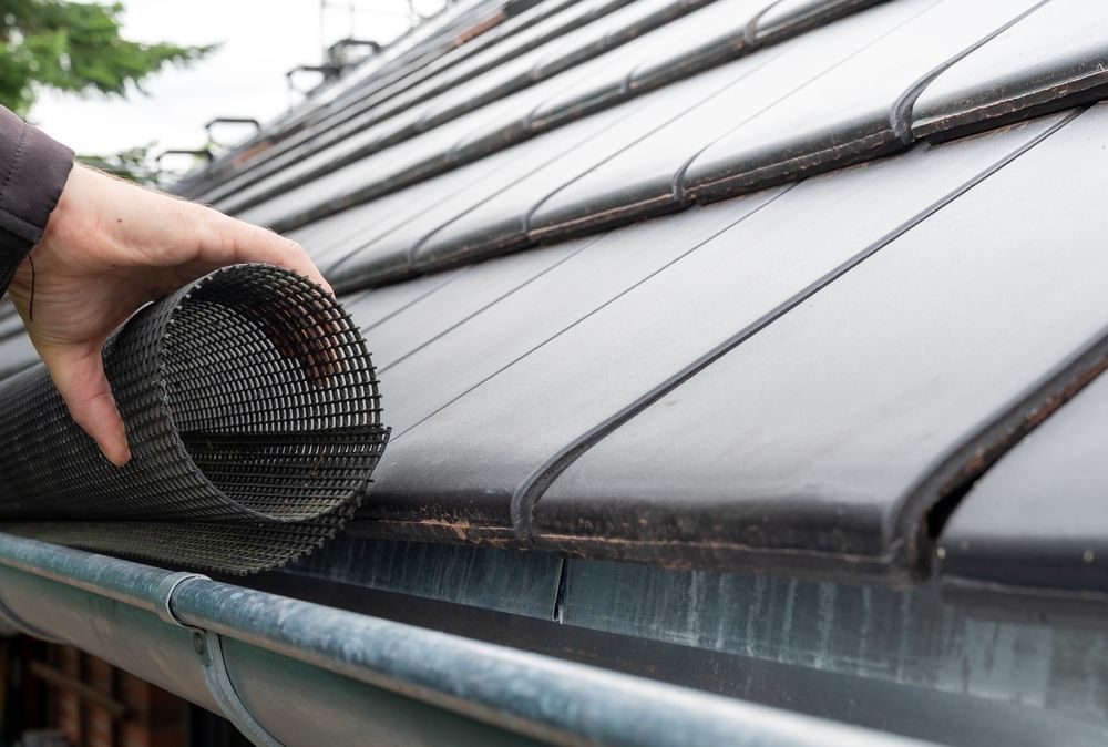 Person installing a black mesh gutter guard on a dark tile roof.